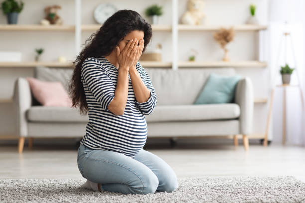 a woman with the baby blues or post partum depression showing signs of sadness while sitting on the floor in a striped blue shirt and blue jeans. 